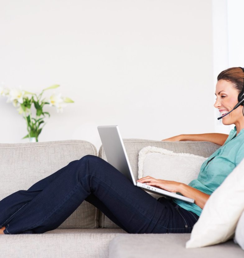 Side view of happy business woman on sofa using laptop and headset at home